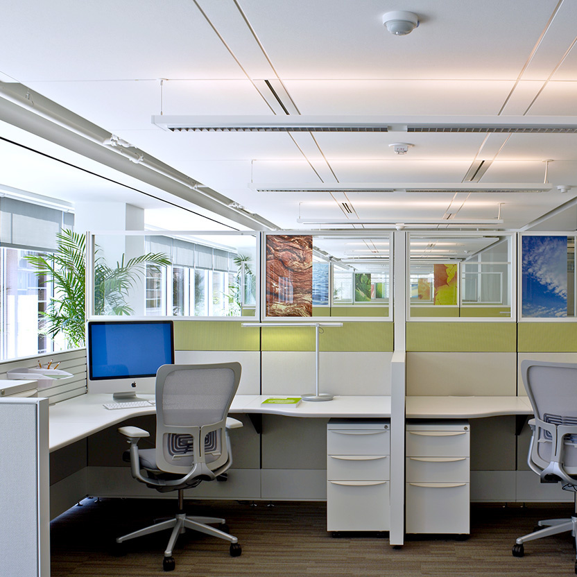 Two cubicles sit in the foreground with two chairs turned into the desks. There is a desktop computer on the left desk, and the office space expands backwards, showcasing natural light coming in from the left and artificial light beams on the ceilings.