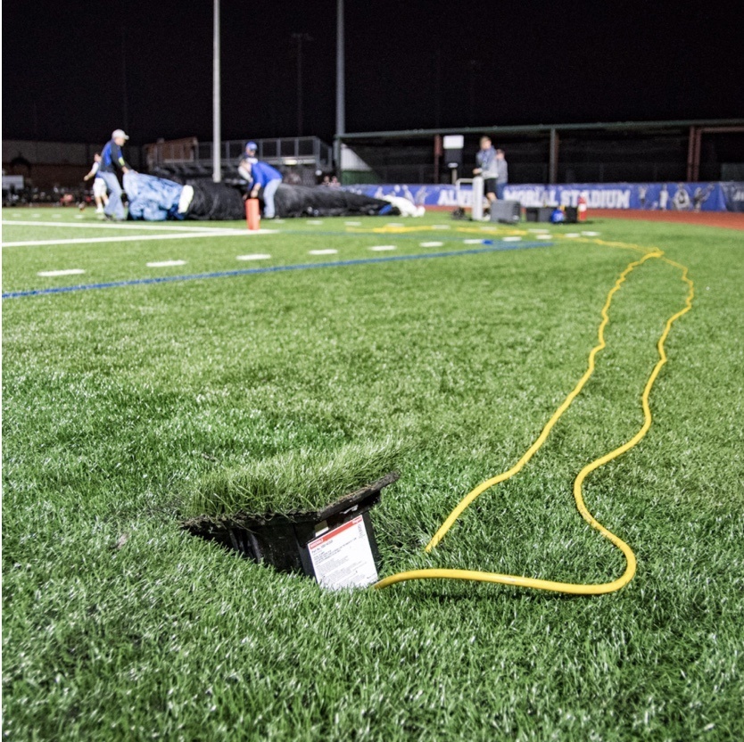 Outdoor ground box in grass with yellow cords emerging and student athletes in background
