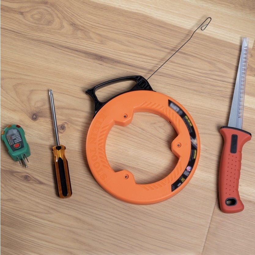Expanded shot of required tools laid on a wooden floor; outlet tester, screwdriver, cable snake reel, drywall saw