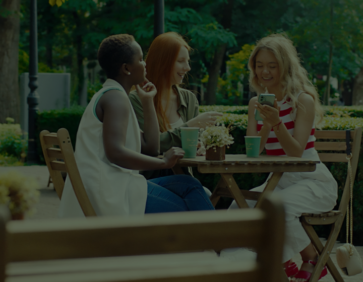 Three woman sitting at outdoor picnic table looking at the phone of the girl sitting on the right